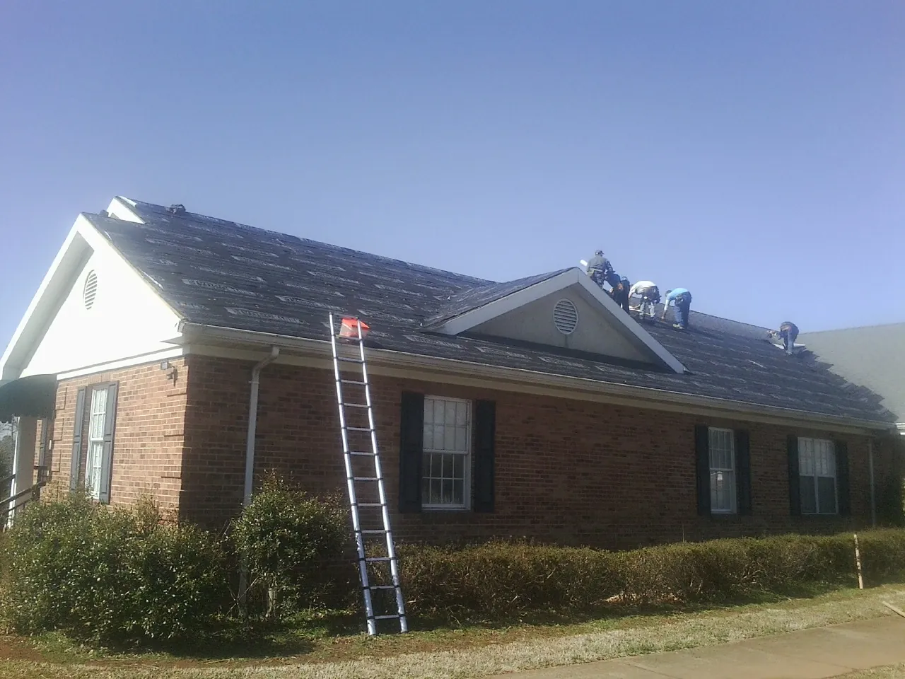 Crew tearing shingles off a brick ranch roof with a ladder set — Metal Roof Repair work by our crew in Erwin
